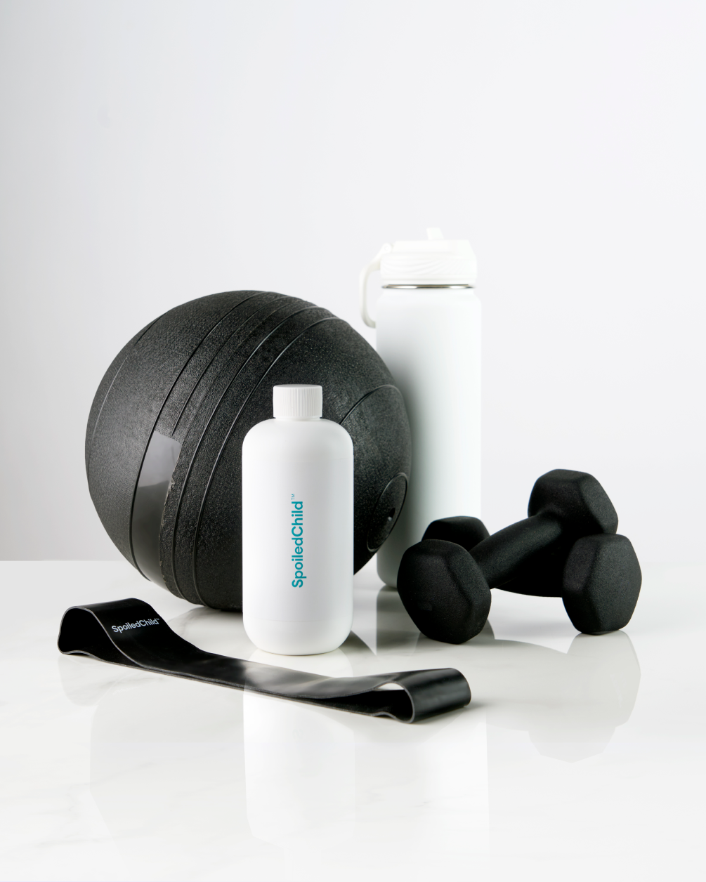 A white SpoiledChild N29 bottle displayed with fitness equipment, including a black medicine ball, two dumbbells, a resistance band, and a white water bottle.