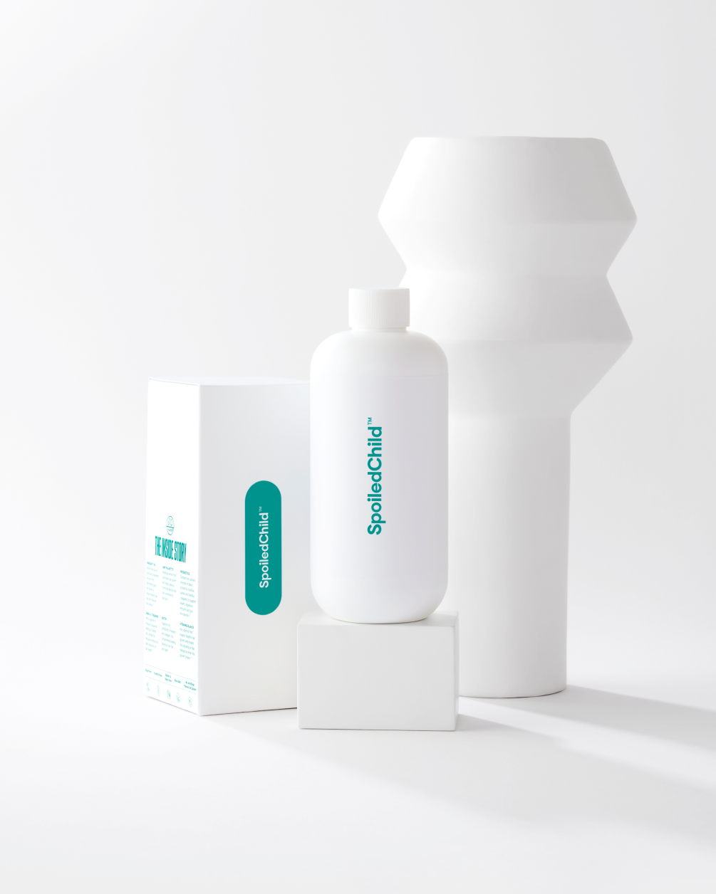 A white SpoiledChild N29 bottle stands on a white block next to its product box and a white sculptural vase.